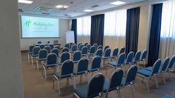 A conference room with rows of blue chairs, a projection screen displaying 'Holiday Inn', and a whiteboard.