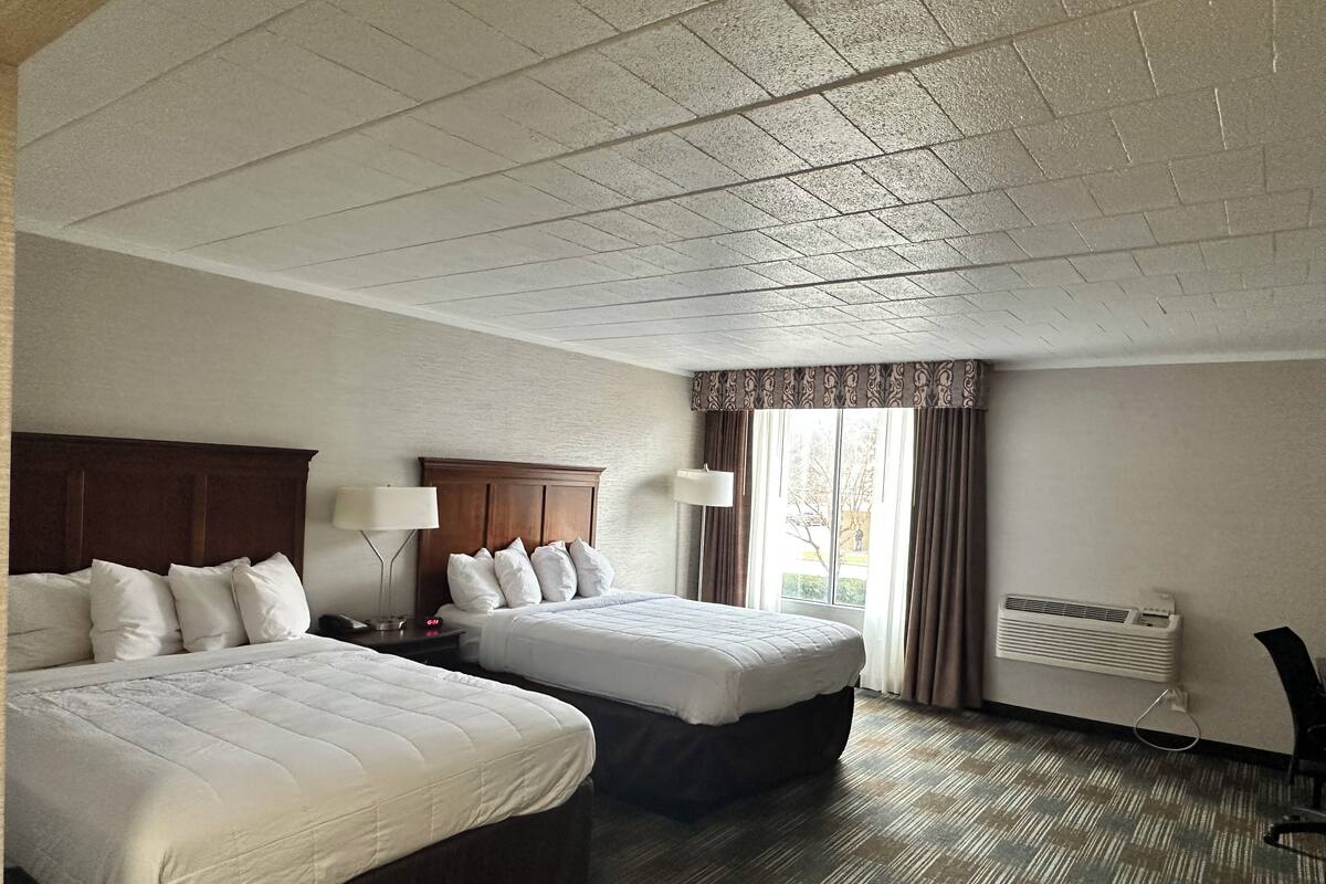 A hotel room with two beds, a large window with curtains, a ceiling fan, and a wall-mounted heater.
