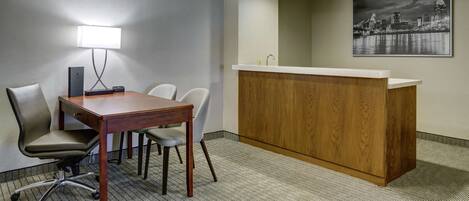 A hotel room with a wooden desk, two chairs, a white desk lamp, a wooden reception desk, and a cityscape painting on the wall.