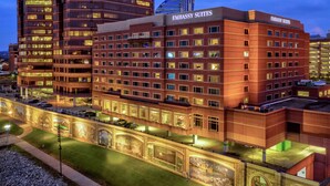 Um hotel com vários andares, com a sinalização "EMBASSY SUITES", apresentando um design arquitetônico moderno, uma entrada destacada e uma área de estacionamento.