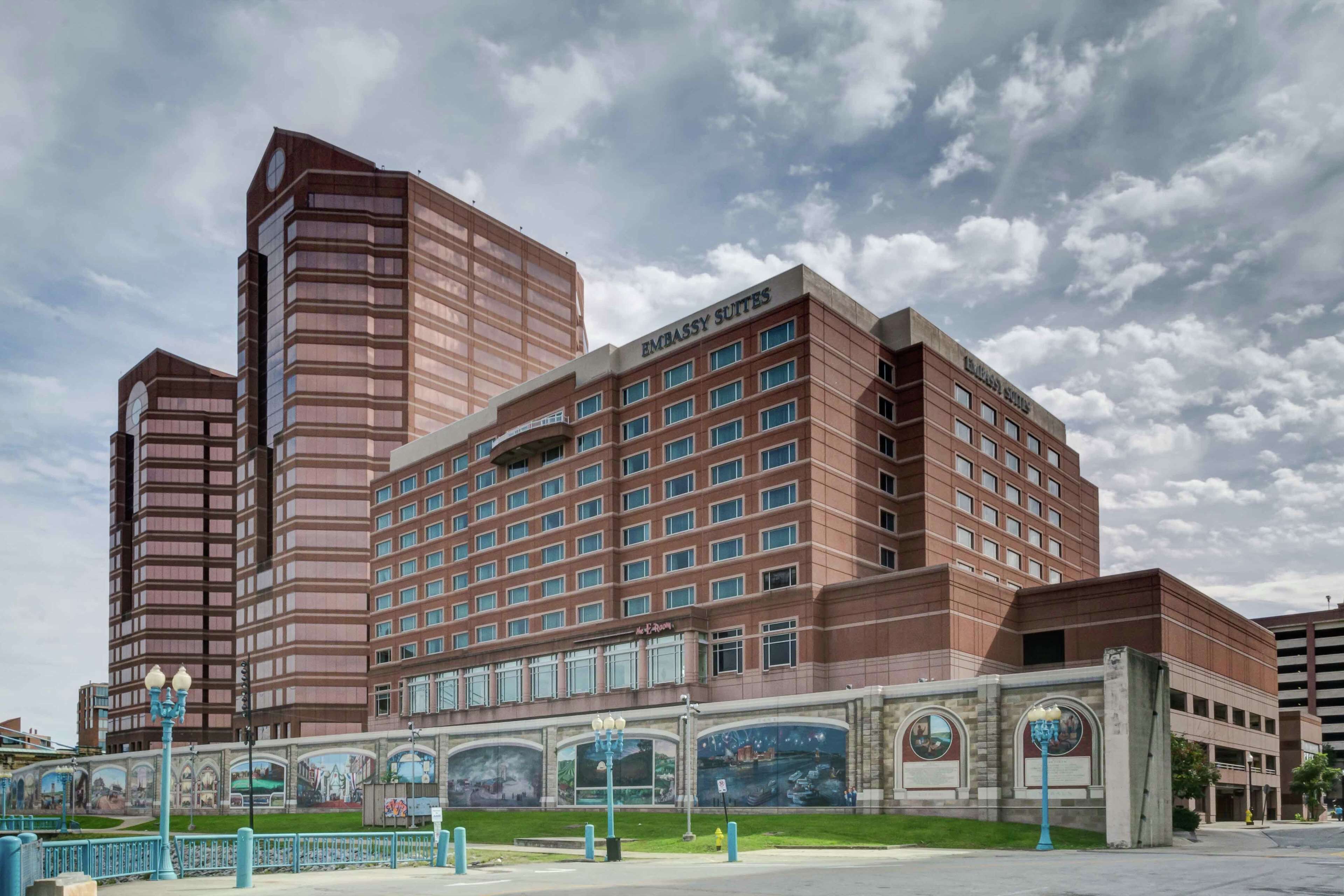 Un hotel de varios pisos con un llamativo letrero de "Embassy Suites", que presenta un diseño arquitectónico moderno con una combinación de fachadas de vidrio y ladrillo.