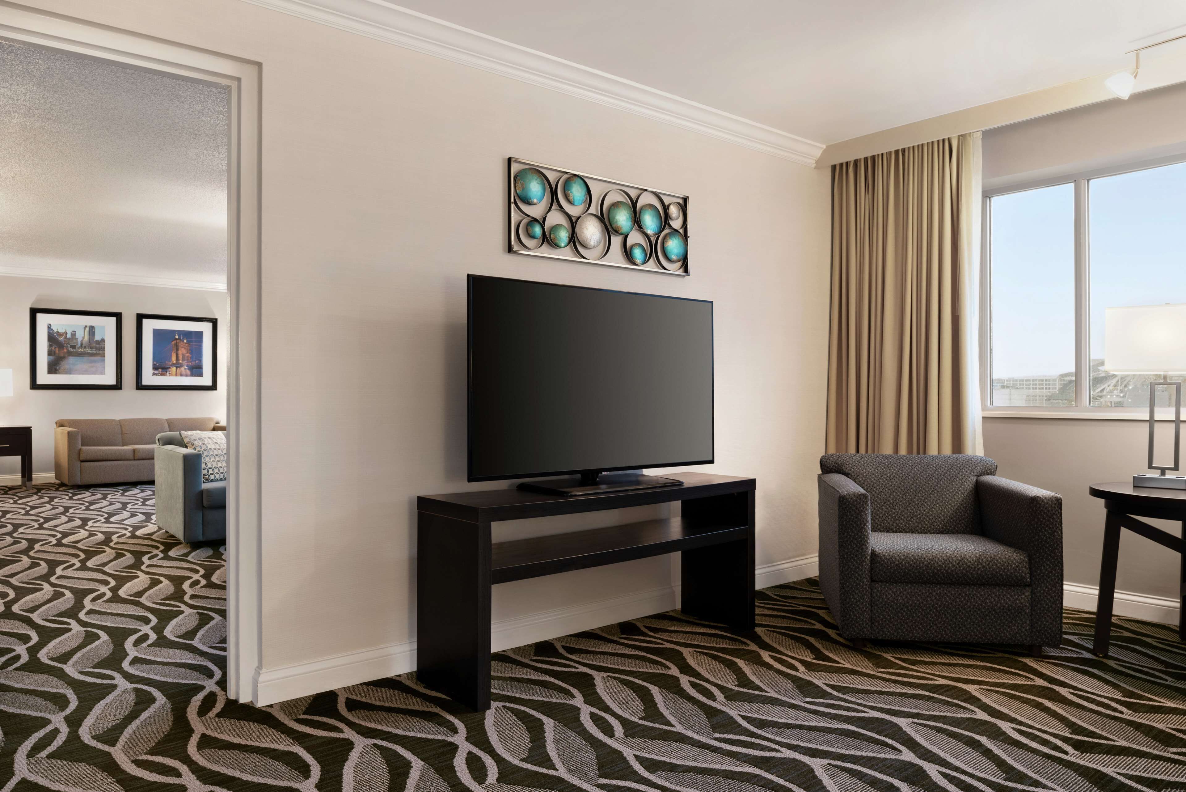 A hotel room with a large flat-screen TV on a black stand, a patterned carpet, a grey armchair, and a window with curtains.
