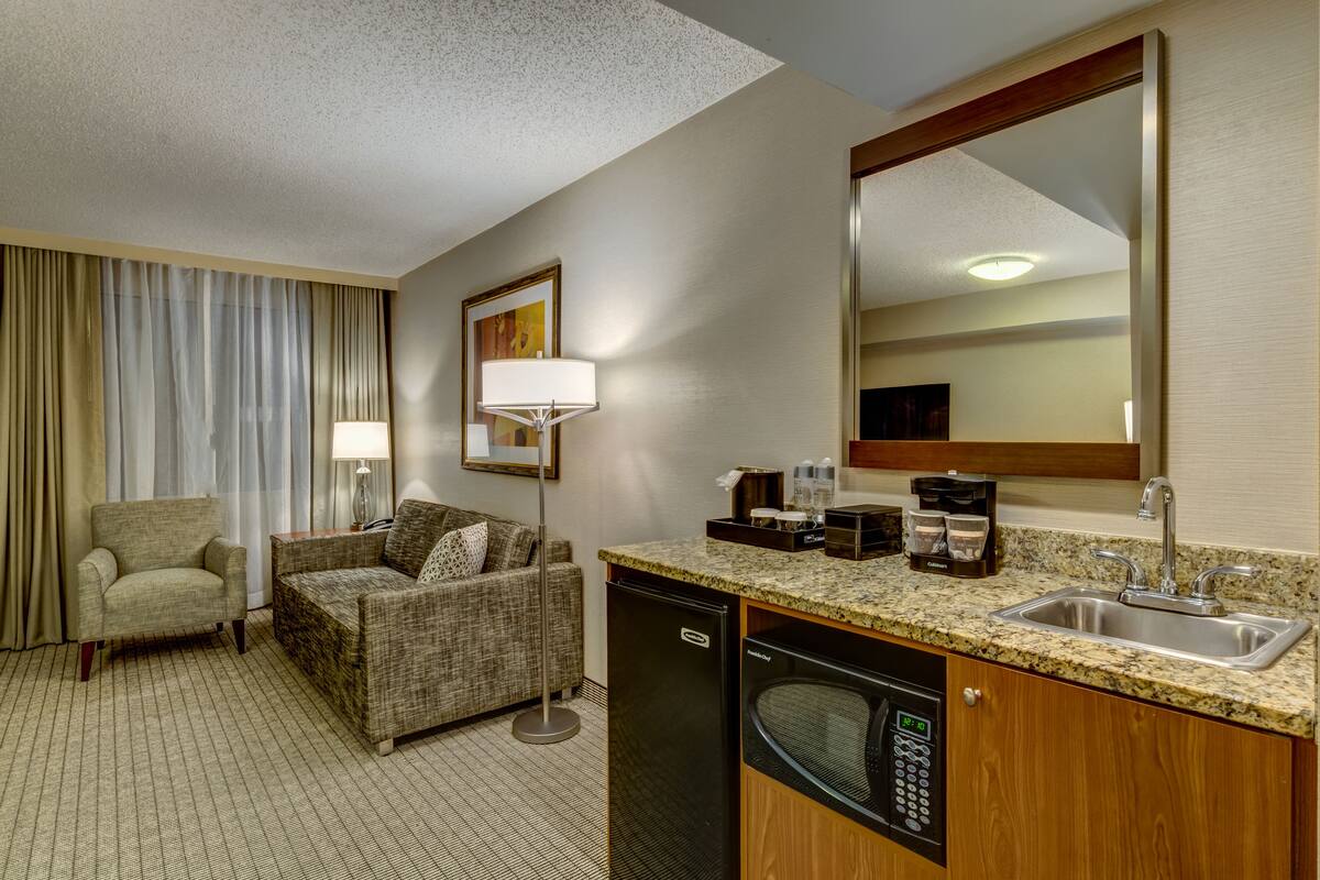 A hotel room with a kitchenette, a sofa, a chair, a large mirror, and a window with curtains.