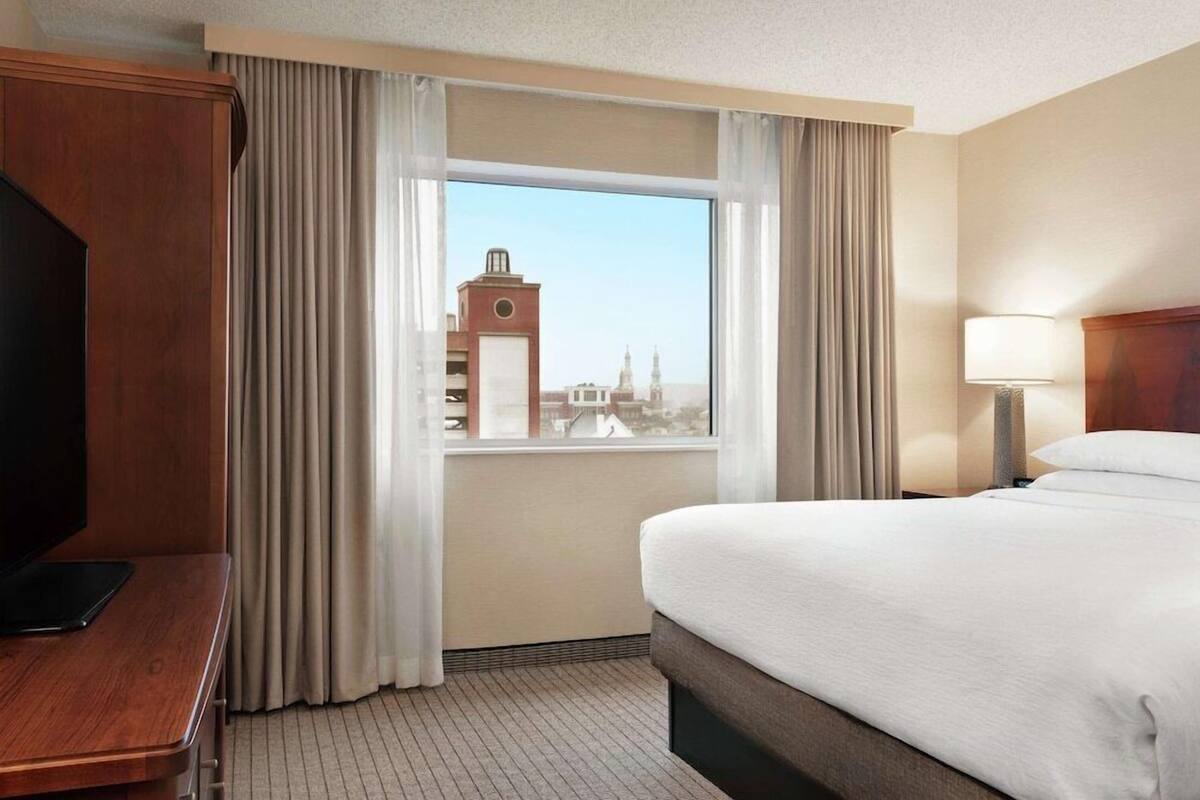 A hotel room with a bed, a desk, a TV, and a window with a cityscape view.