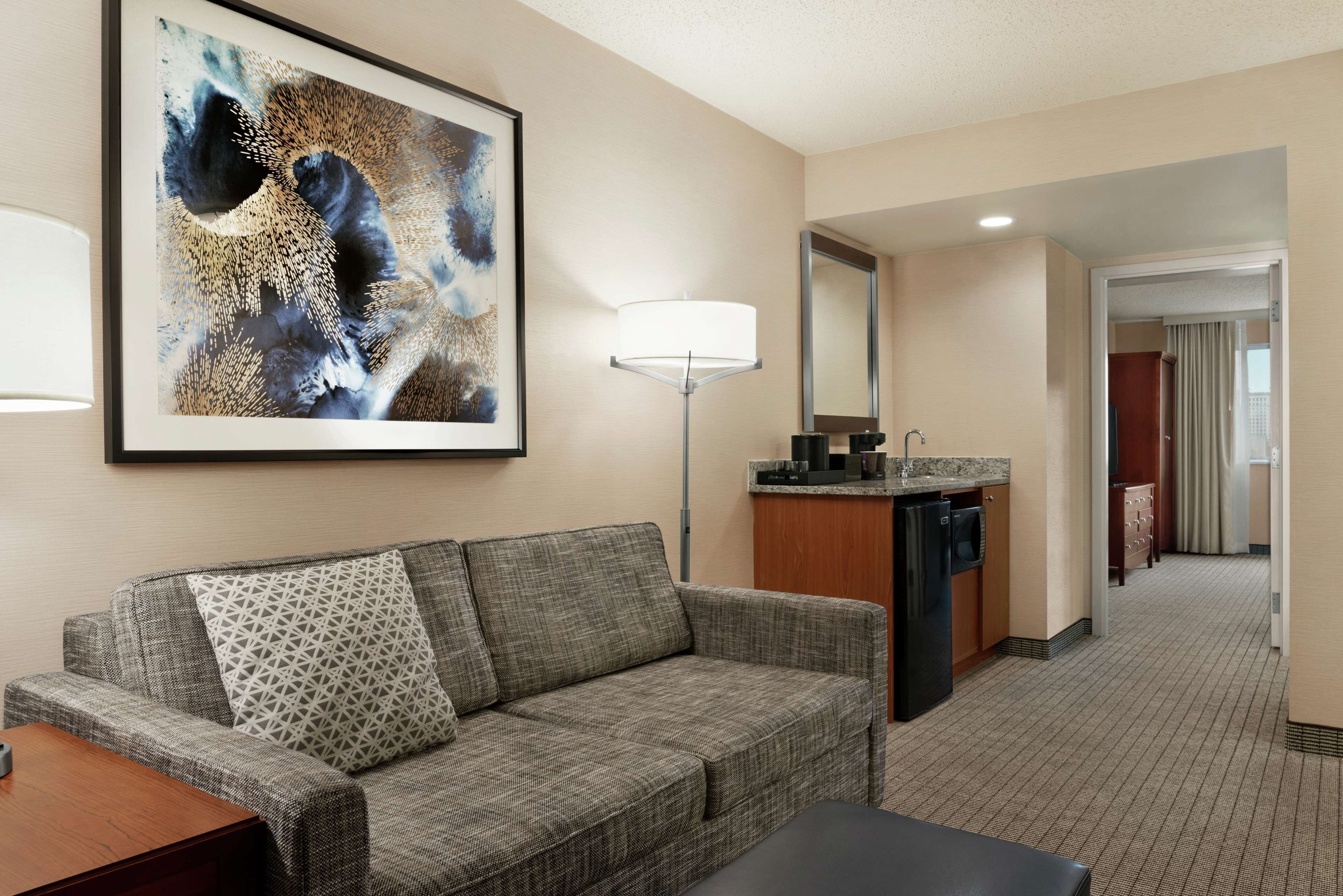 A hotel room with a grey sofa, a patterned pillow, a framed abstract painting, a floor lamp, a kitchen area with a refrigerator, and a mirror.