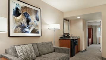 A hotel room with a grey sofa, a patterned pillow, a framed abstract painting, a floor lamp, a kitchen area with a refrigerator, and a mirror.