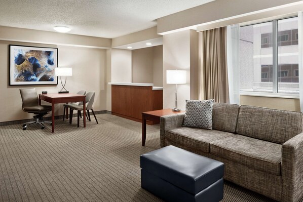 A hotel room with a sofa, a desk with two chairs, a small table, a lamp, a large window with curtains, and a framed artwork on the wall.