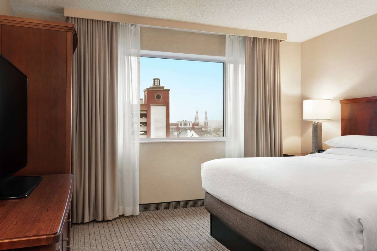 A hotel room with a large bed, a TV, a desk, and a window with a cityscape view.