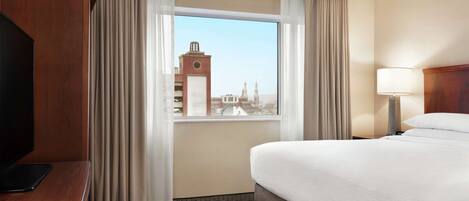 A hotel room with a large bed, a TV, a desk, and a window with a cityscape view.