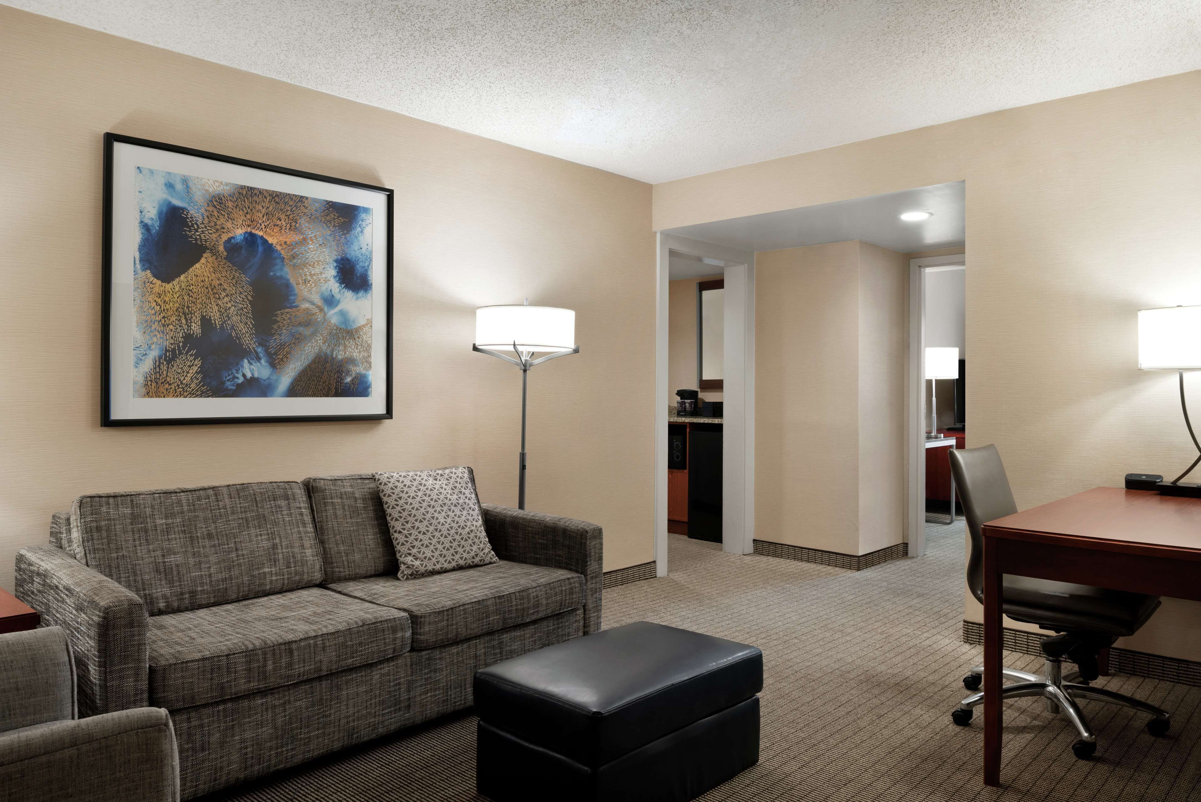 A hotel room with a grey sofa, a black ottoman, a desk with a chair, a floor lamp, and a framed abstract painting on the wall.