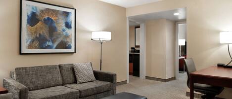 A hotel room with a grey sofa, a black ottoman, a desk with a chair, a floor lamp, and a framed abstract painting on the wall.