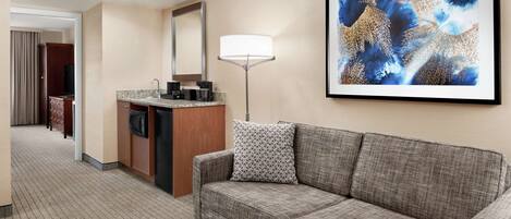 A hotel room with a kitchenette, a sofa, a flat-screen TV, a large abstract painting, and a floor lamp.