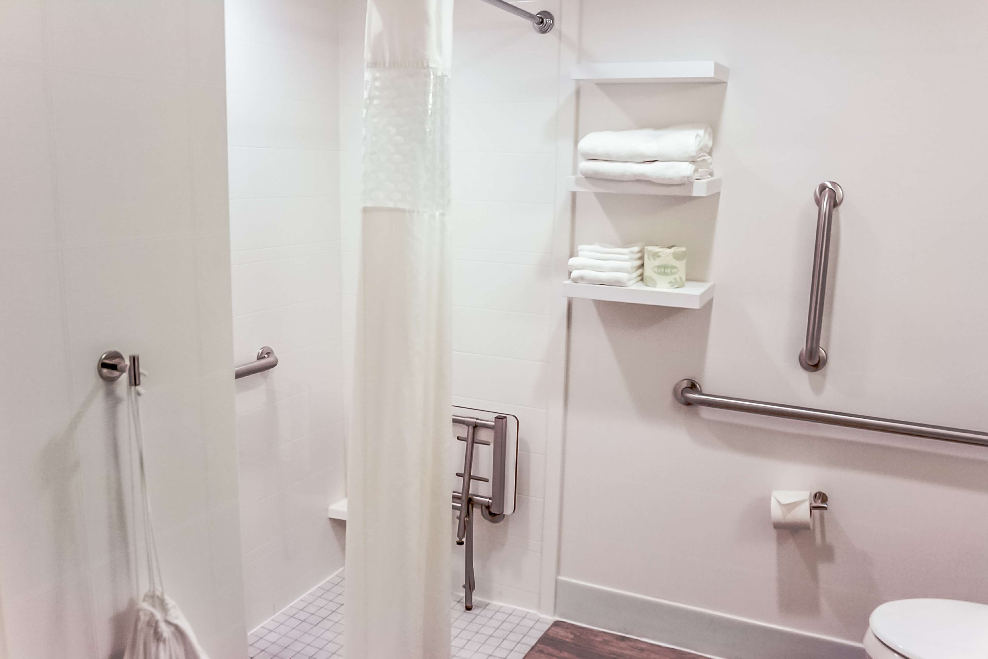 Une salle de bain équipée d’une douche, de barres d’appui, de toilettes et d’étagères où sont rangées les serviettes.