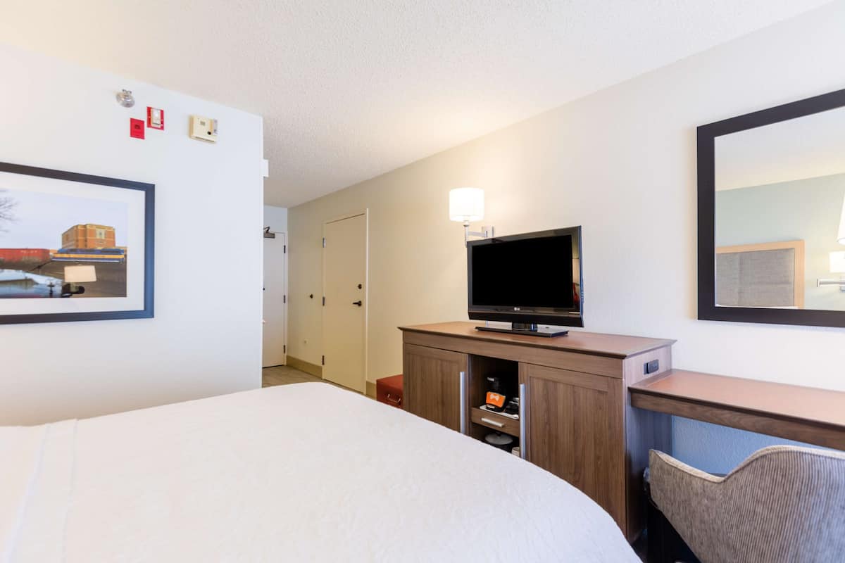 A hotel room with a bed, a desk with a television, a mirror, and a framed picture on the wall.