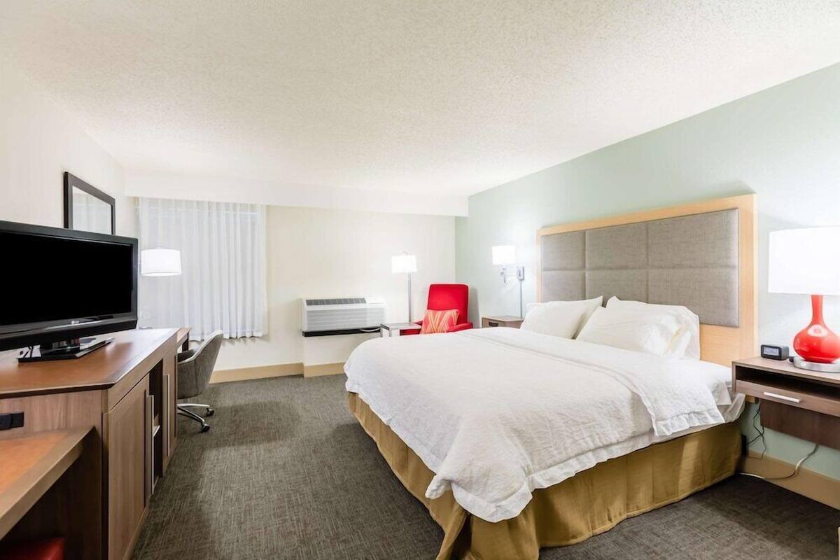 A hotel room with a bed, a desk with a TV, a red chair, and a lamp.