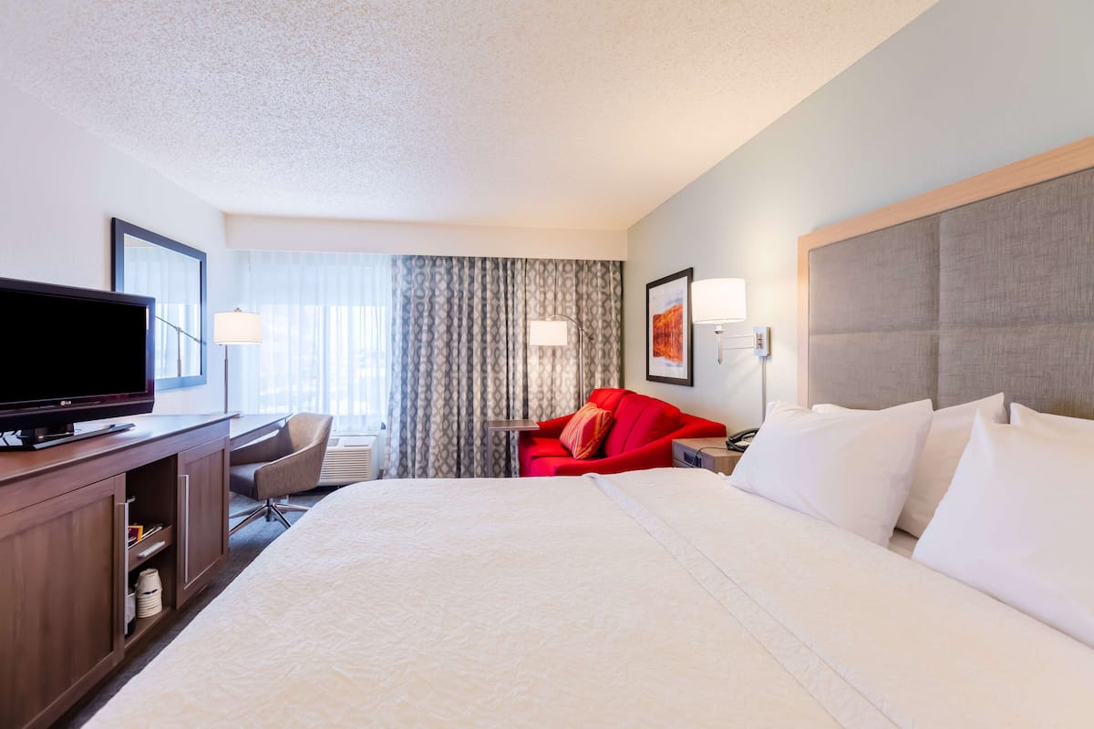 A hotel room with a large bed, a television, a desk with a chair, a red sofa, and a large window with curtains.