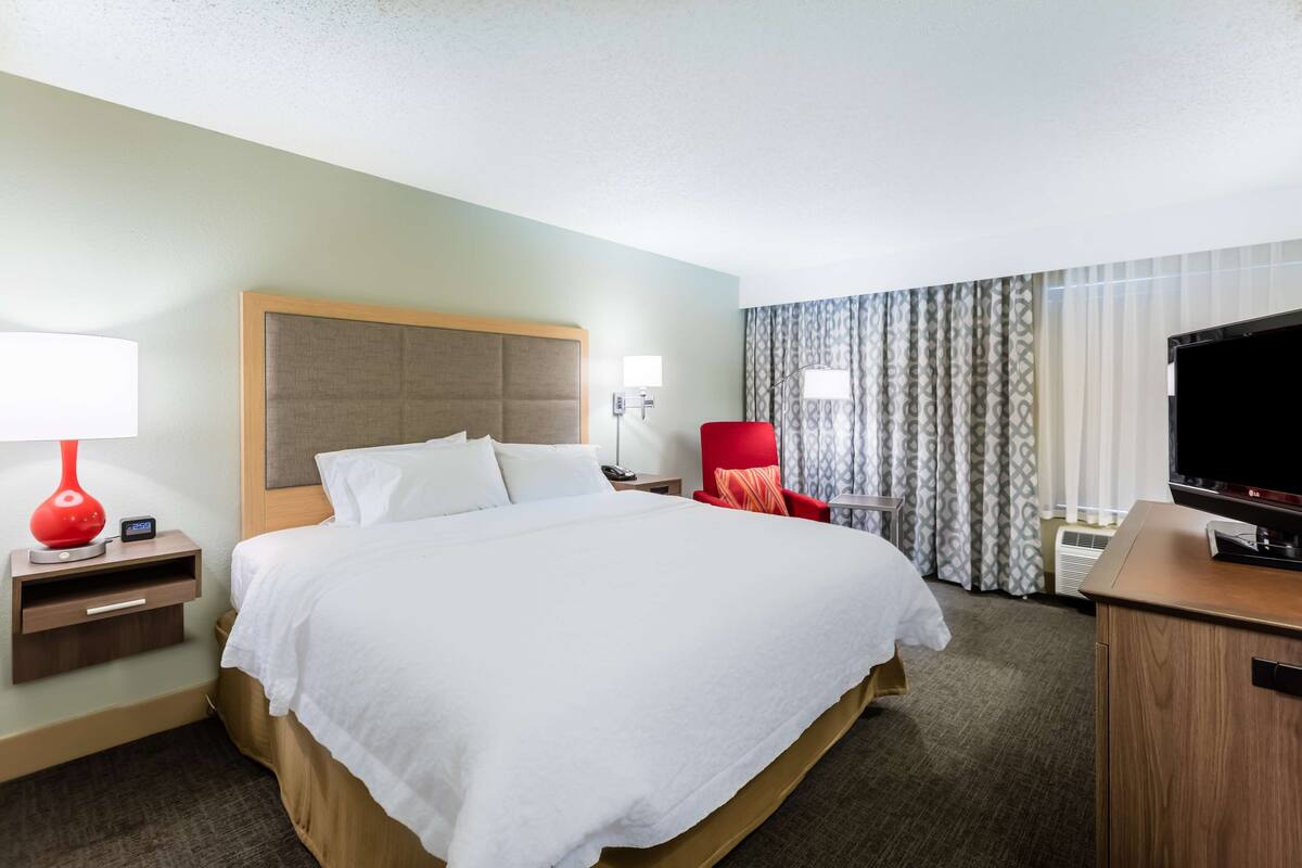 A hotel room with a large bed, a desk with a TV, a red chair, and a nightstand with a lamp.