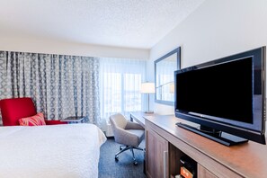 A hotel room with a large bed, a red armchair, a grey office chair, a TV on a wooden stand, and a window with patterned curtains.