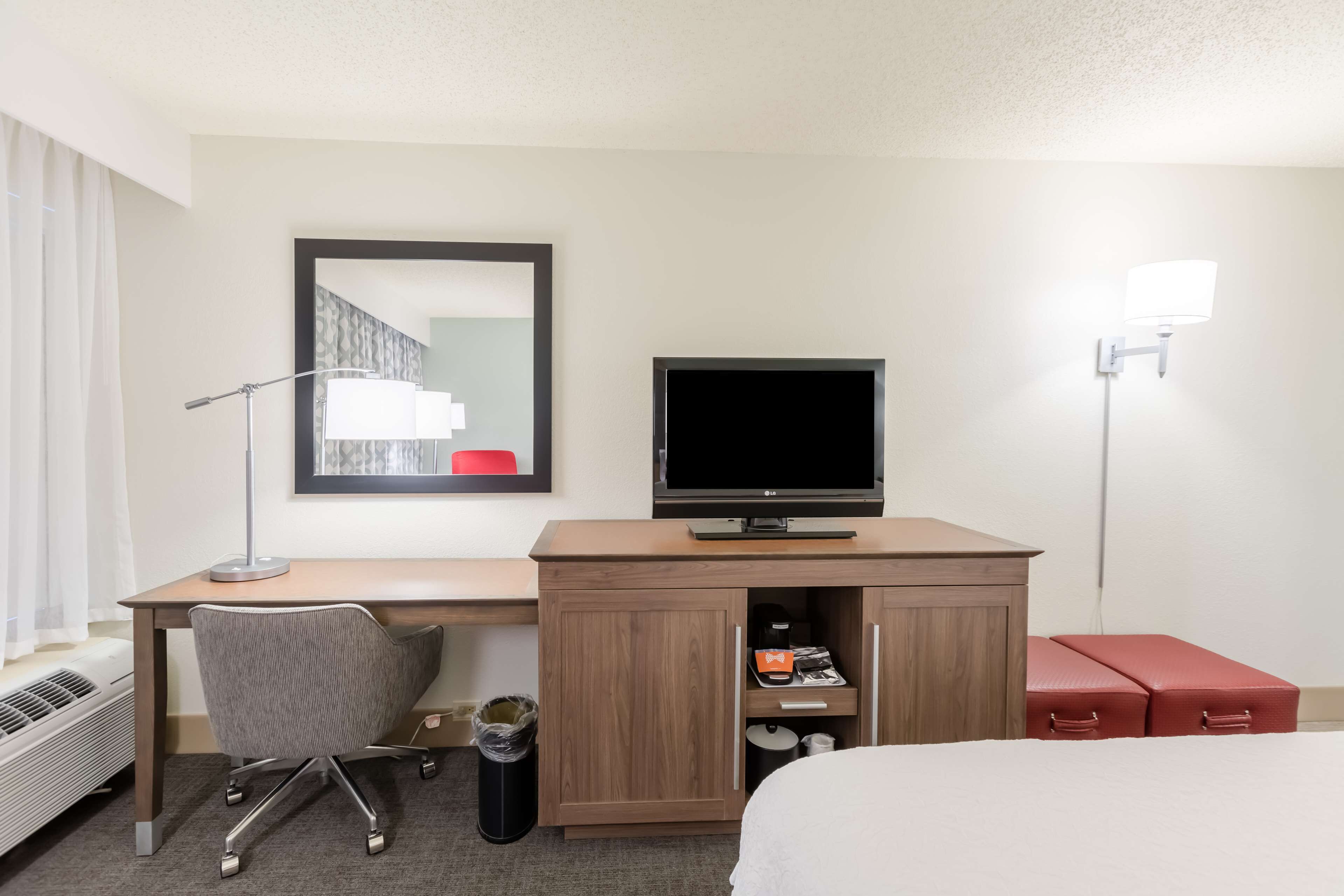 Habitación de hotel con escritorio de madera, televisor, silla, cama, cubo de basura y lámpara.