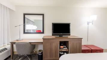 Habitación de hotel con escritorio de madera, televisor, silla, cama, cubo de basura y lámpara.