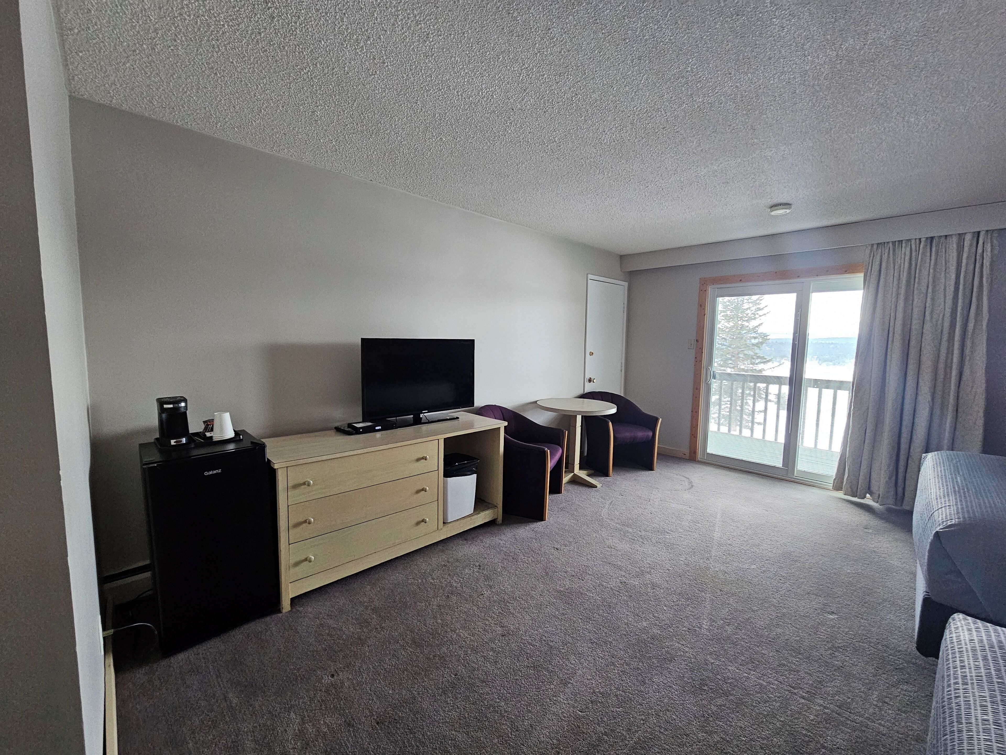 A hotel room with a flat-screen TV on a dresser, a small round table with two purple chairs, a sofa, and a sliding glass door to the outside.