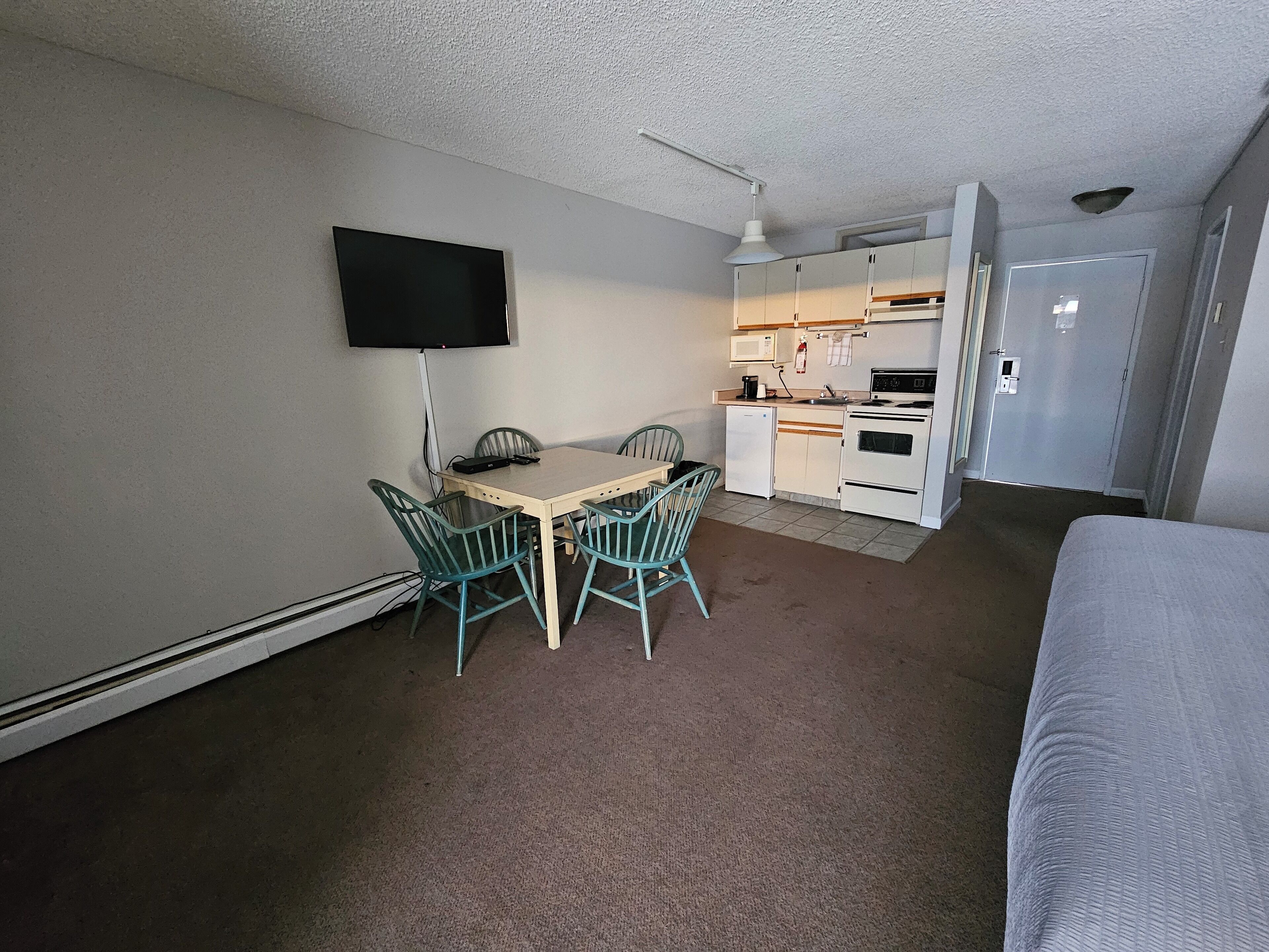 A compact hotel room with a single bed, a small kitchenette, a dining area with a table and chairs, and a TV mounted on the wall.