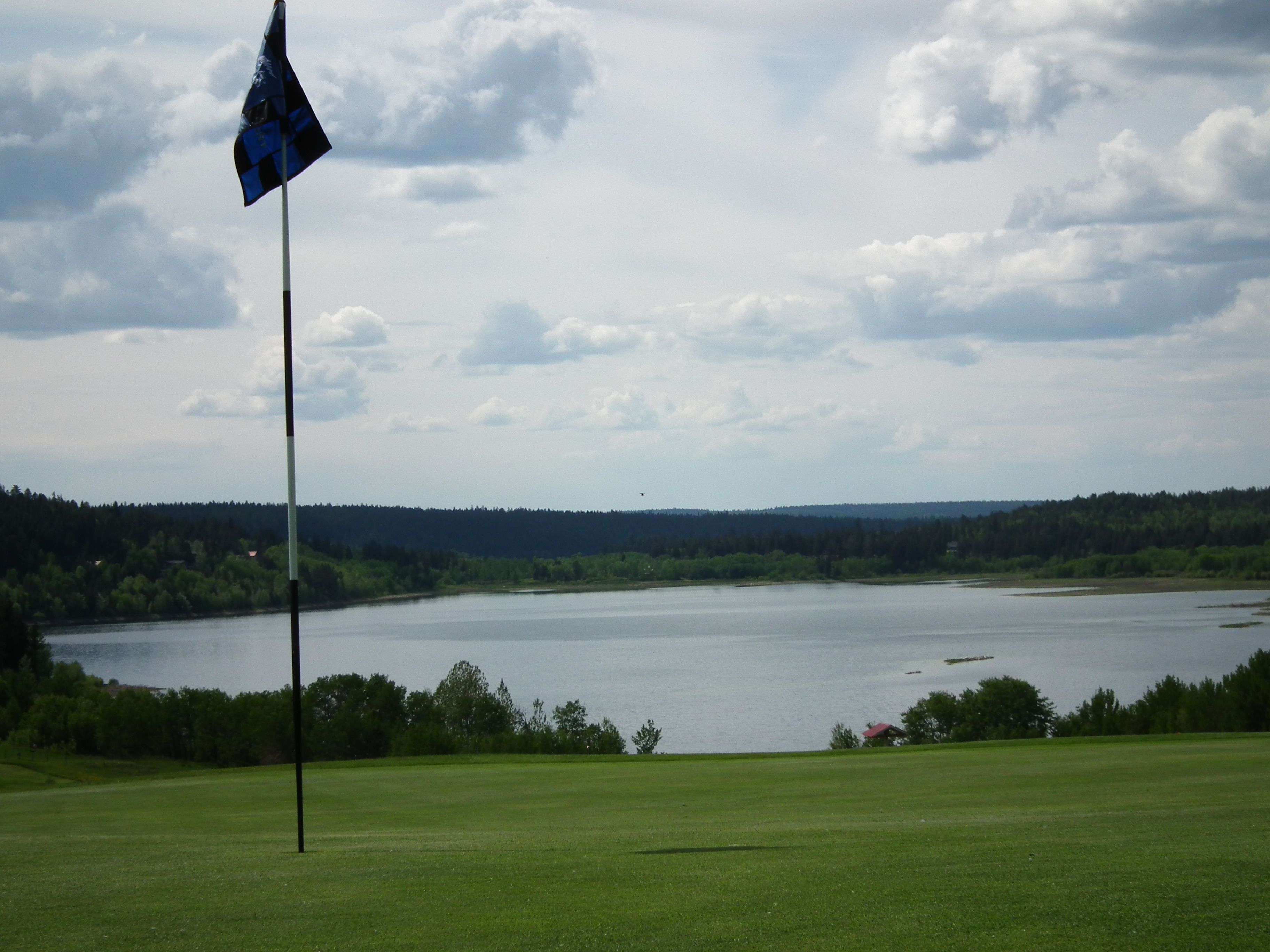 Un terrain de golf avec un drapeau sur un mât, une étendue d’eau et un paysage boisé en arrière-plan.