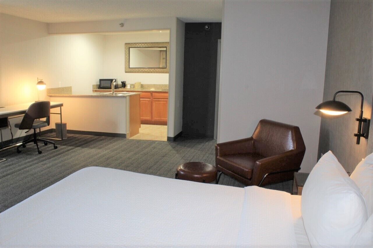 A hotel room with a bed, a desk, a chair, a small stool, and a kitchenette.