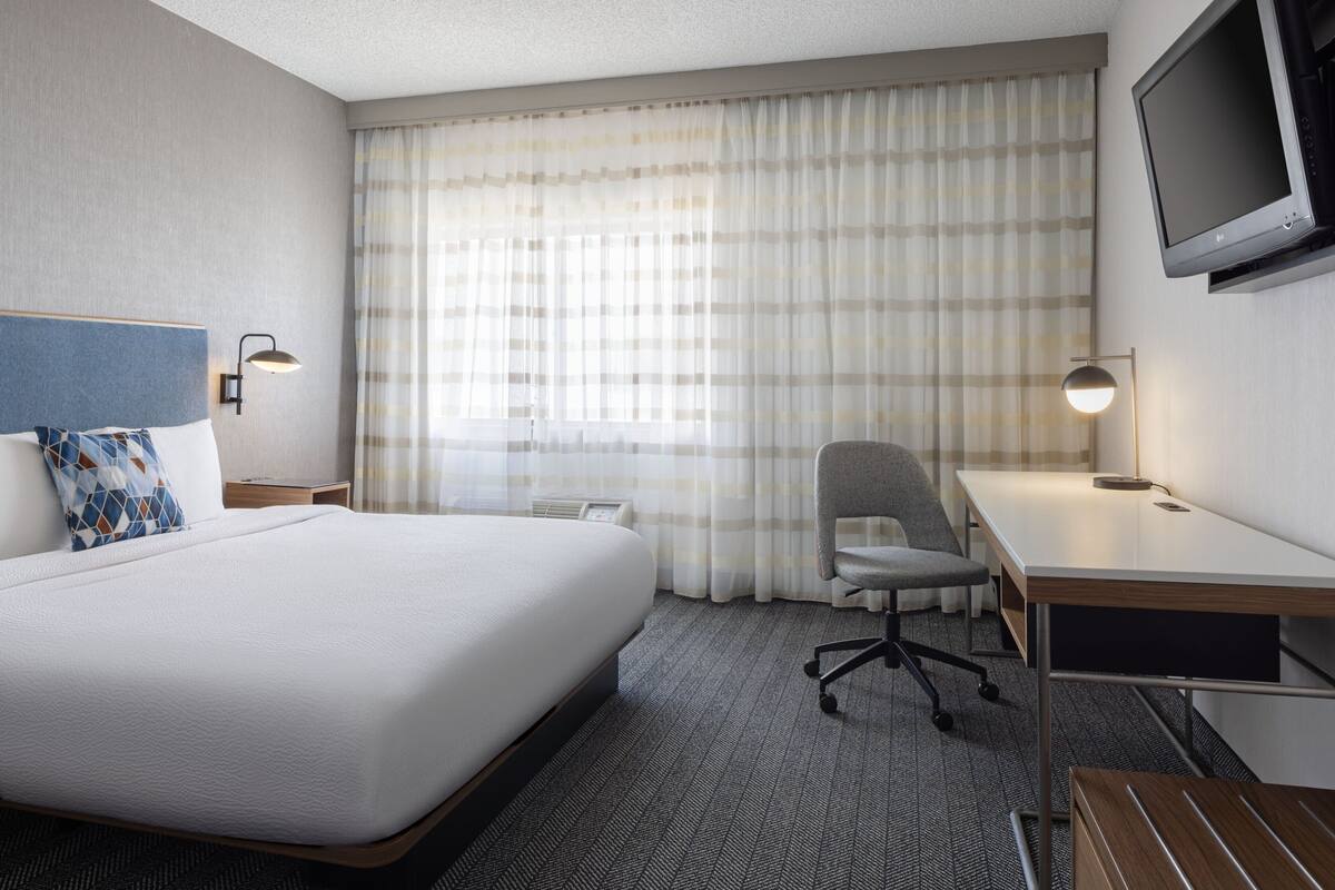 A hotel room with a large bed, a desk, a chair, a TV, and a window with curtains.
