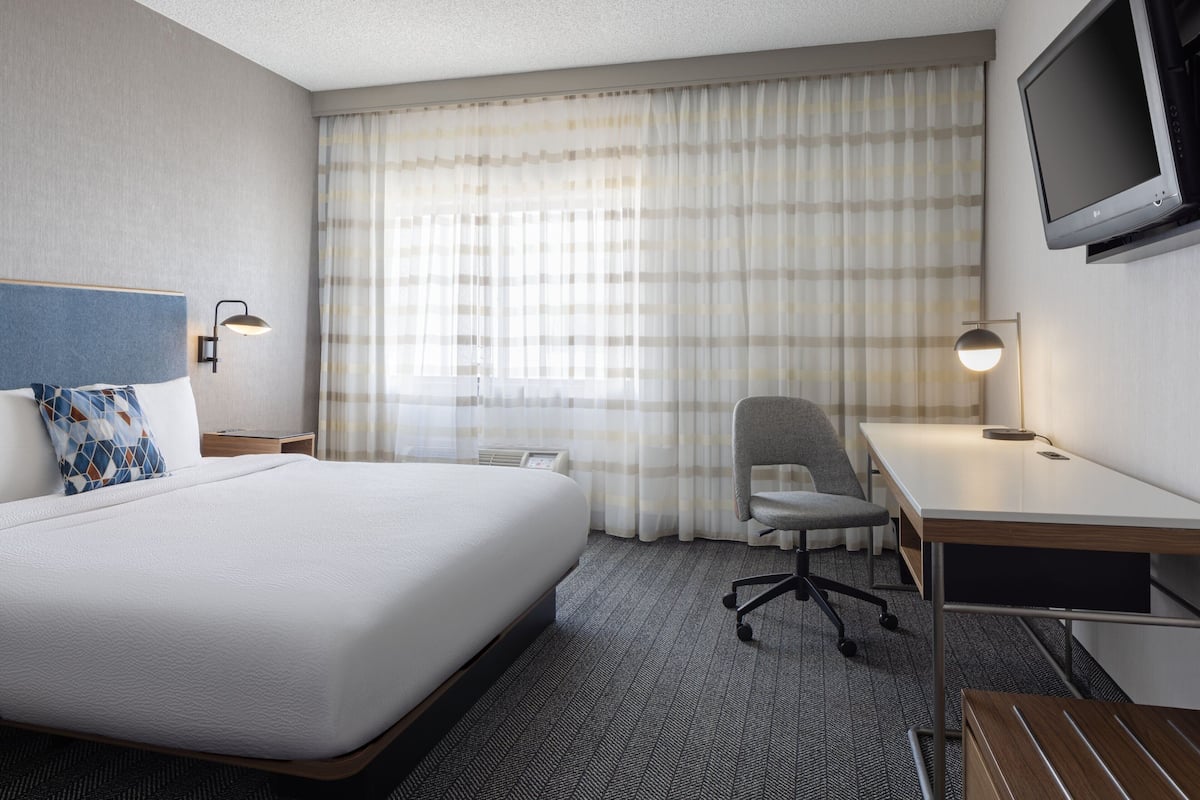 Quarto de hotel com uma cama grande, uma escrivaninha, uma cadeira, uma televisão e uma janela com cortinas.