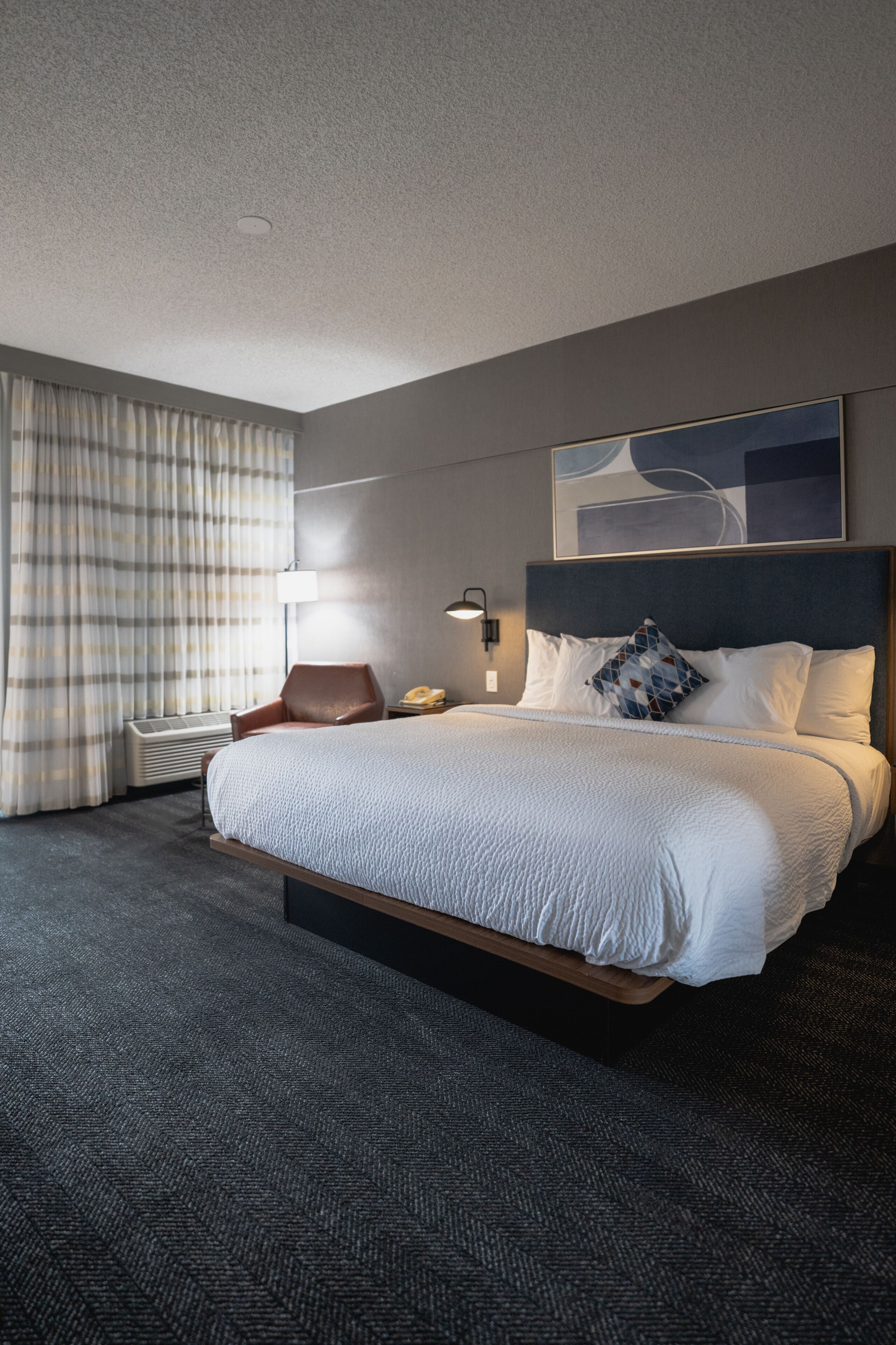 A hotel room with a large bed, a chair, a desk with a lamp, and a window with curtains.