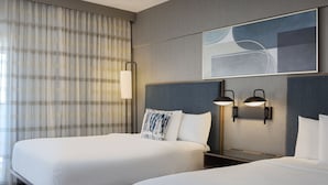 A hotel room with two beds, a large window with curtains, a wall-mounted artwork, and two wall sconces.