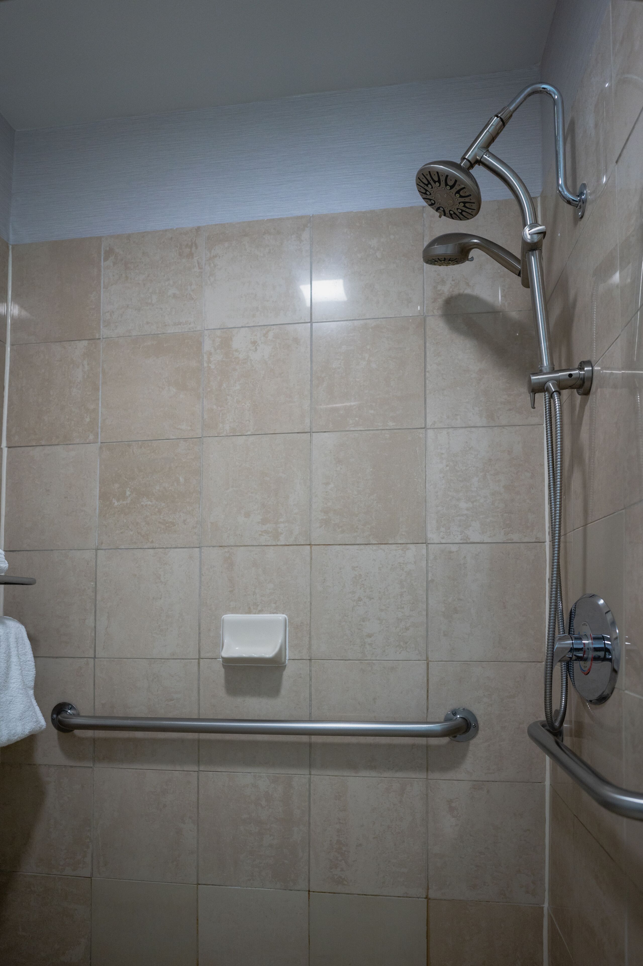 A bathroom with beige tiles, a handheld showerhead, a handheld shower attachment, a grab bar, and a towel rack.
