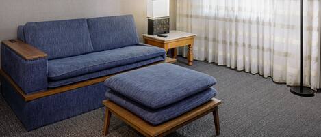 A room with a blue sofa, a matching ottoman, a small wooden side table with a lamp, and a curtain with a checkered pattern.