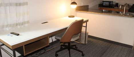 A hotel room with a desk, chair, and a small kitchenette area.