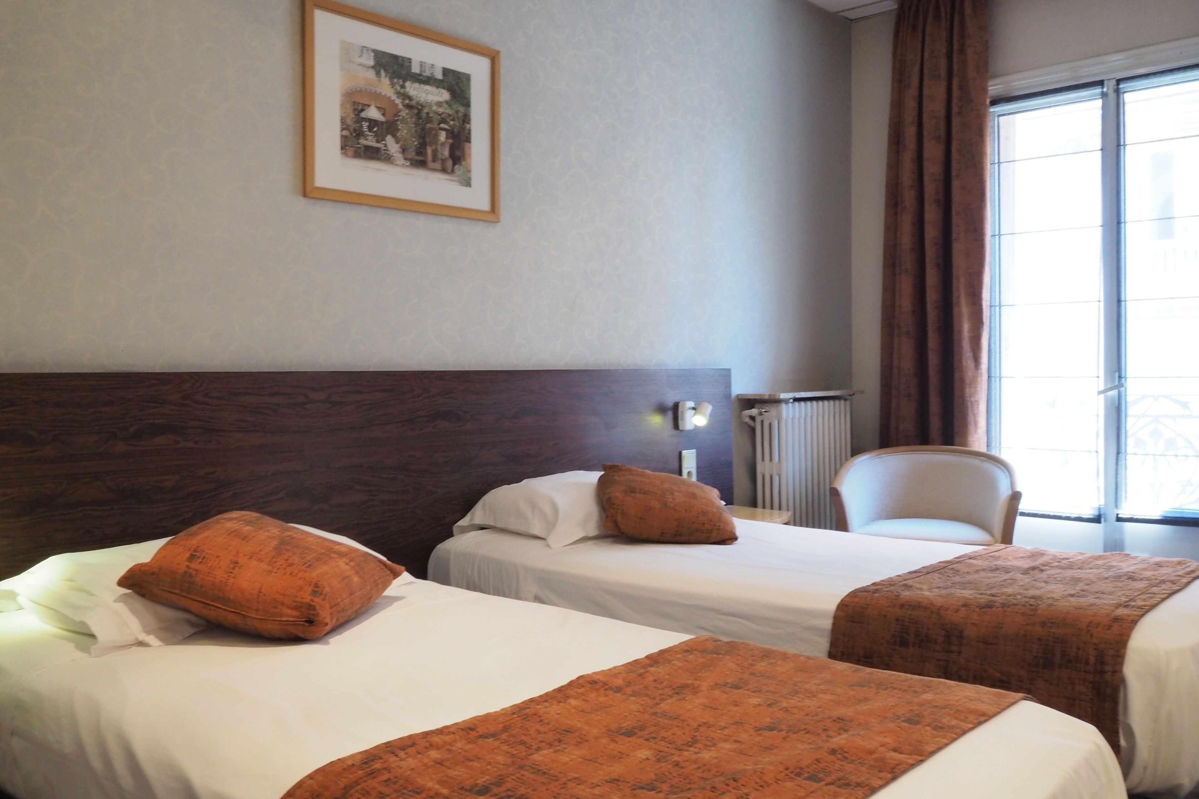 A hotel room with two beds, a wooden headboard, a framed picture on the wall, a chair, and a window with curtains.
