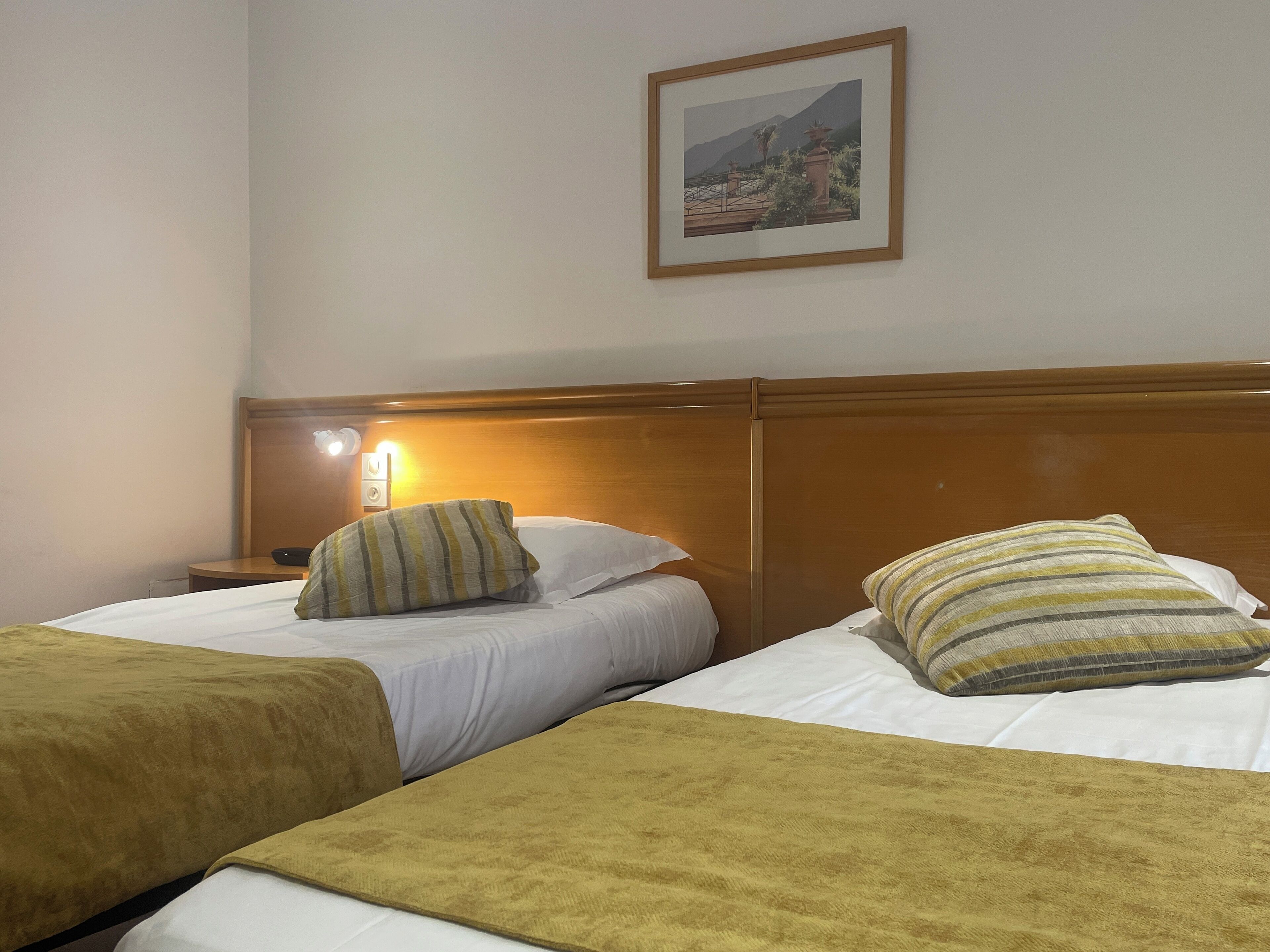 A hotel room with two beds, each with a wooden headboard, white linens, and striped pillows. A framed picture hangs on the wall above the beds.