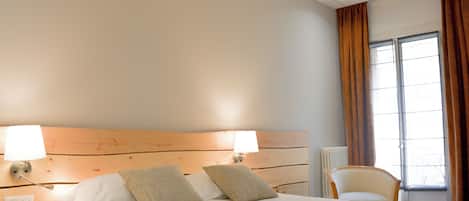 Habitación de hotel con una cama grande, dos lámparas de noche, un cabecero de madera, una ventana con cortinas y una silla.