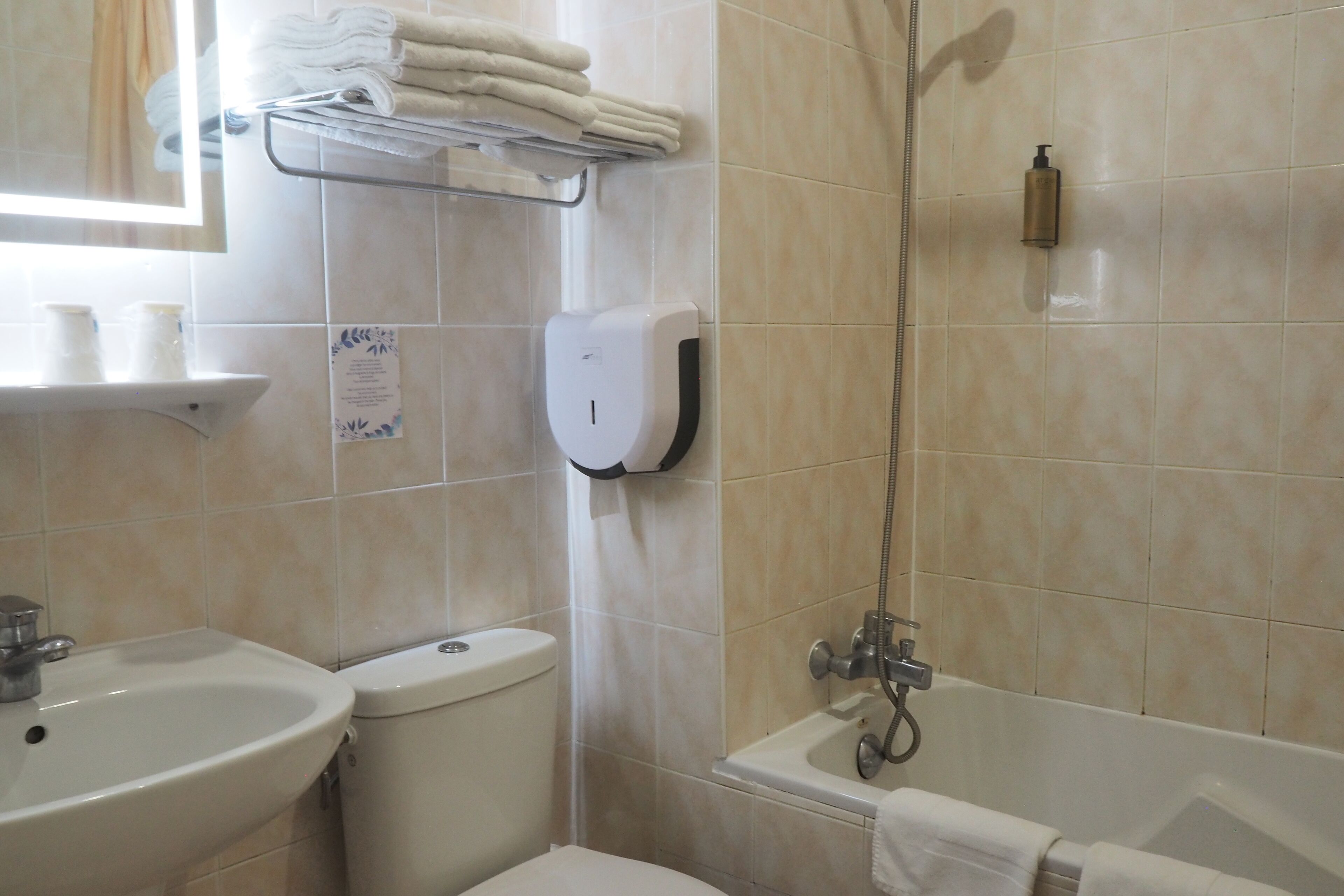 A bathroom with a sink, toilet, and bathtub. There are towels stacked on a shelf and a soap dispenser mounted on the wall.