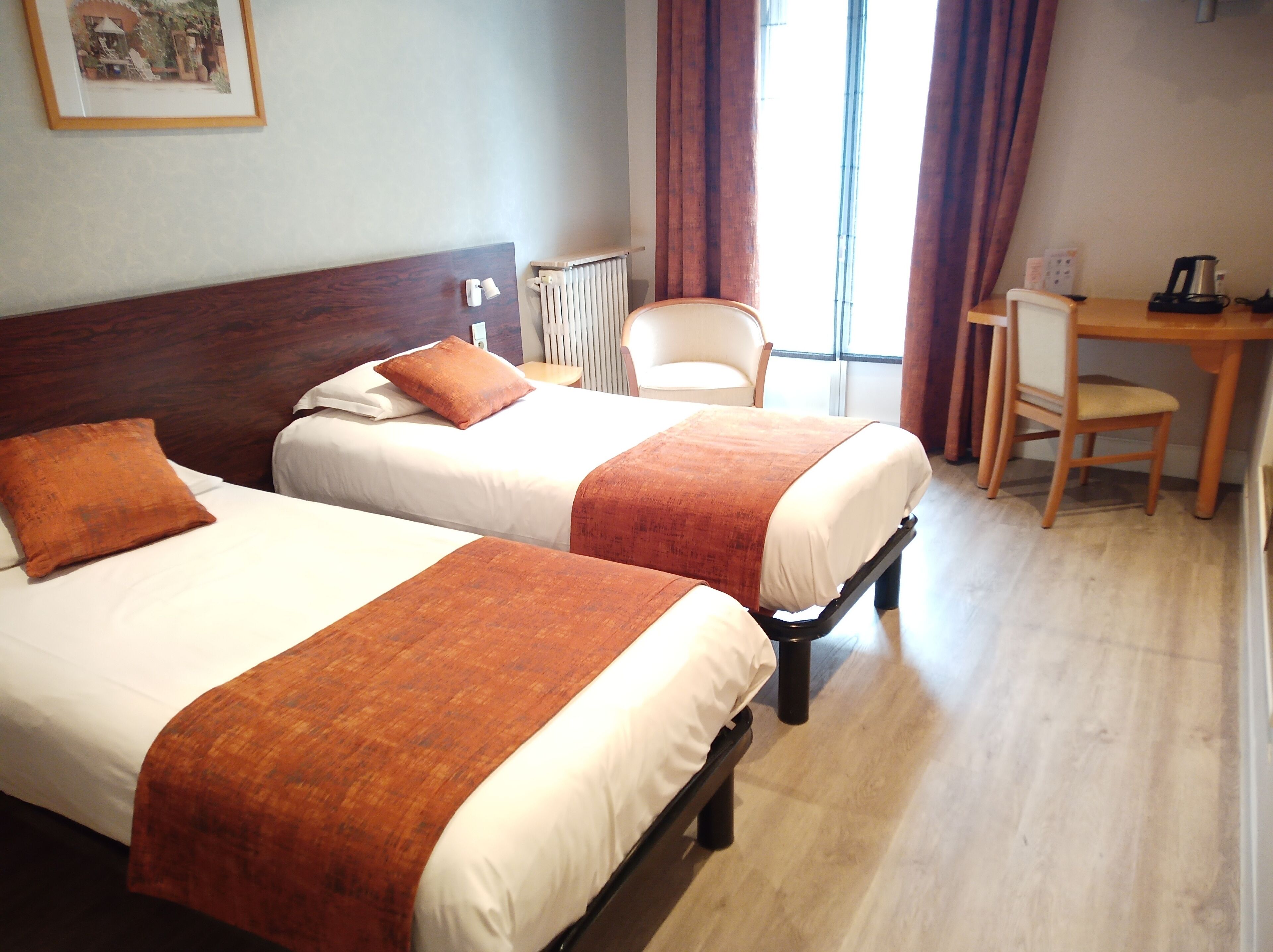 Ein Hotelzimmer mit zwei Betten, einem hölzernen Kopfteil, einem Heizkörper, einem Sessel, einem Schreibtisch und einem Fenster mit Vorhängen.