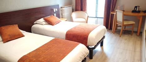Habitación de hotel con dos camas, cabecero de madera, radiador, una silla, un escritorio y una ventana con cortinas.