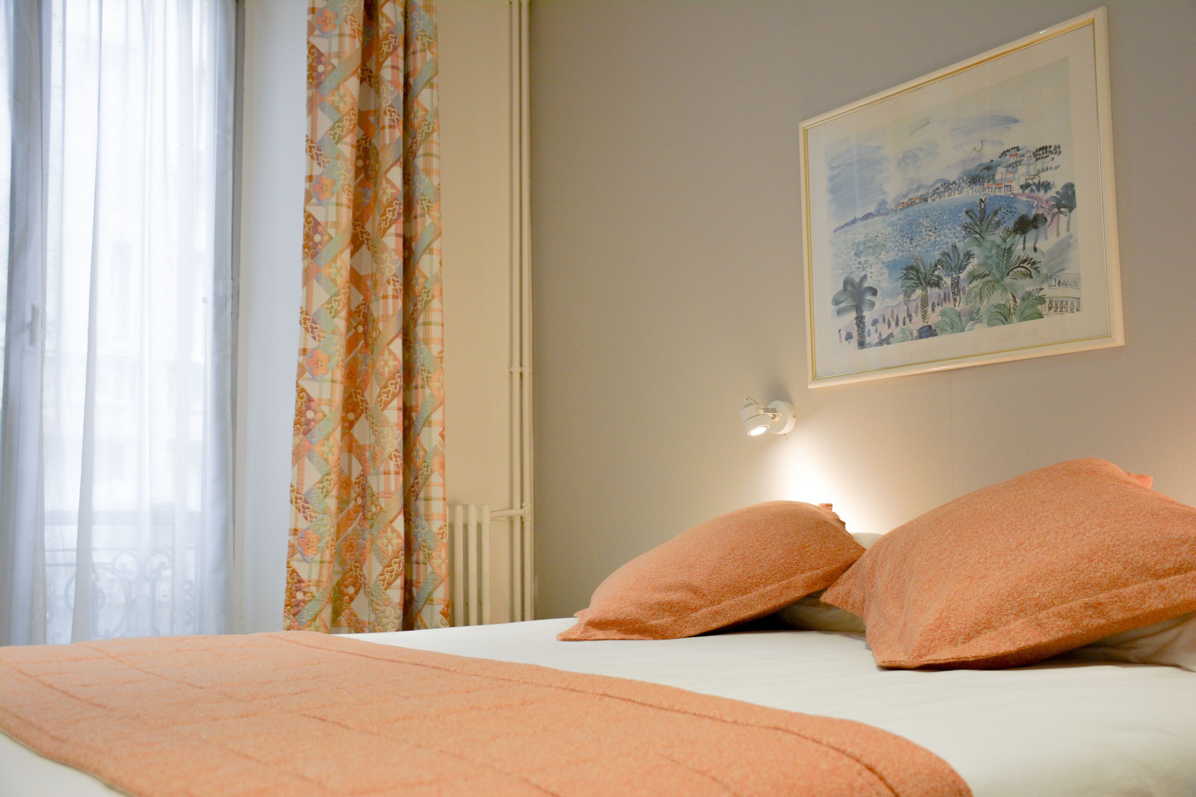 Un dormitorio con cama, ropa de cama naranja, una ventana con cortinas traslúcidas y un cuadro en la pared.