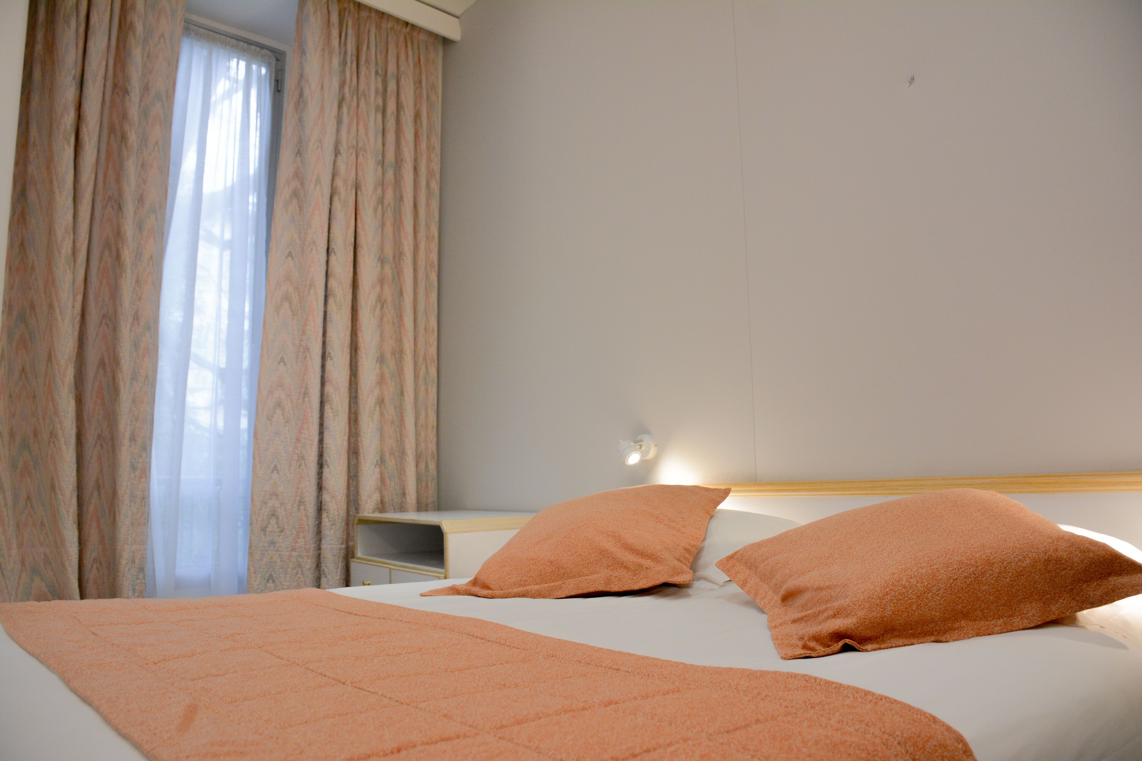 Una cama bien hecha con dos almohadas y una manta, una mesita de noche con lámpara, y una ventana con cortinas.