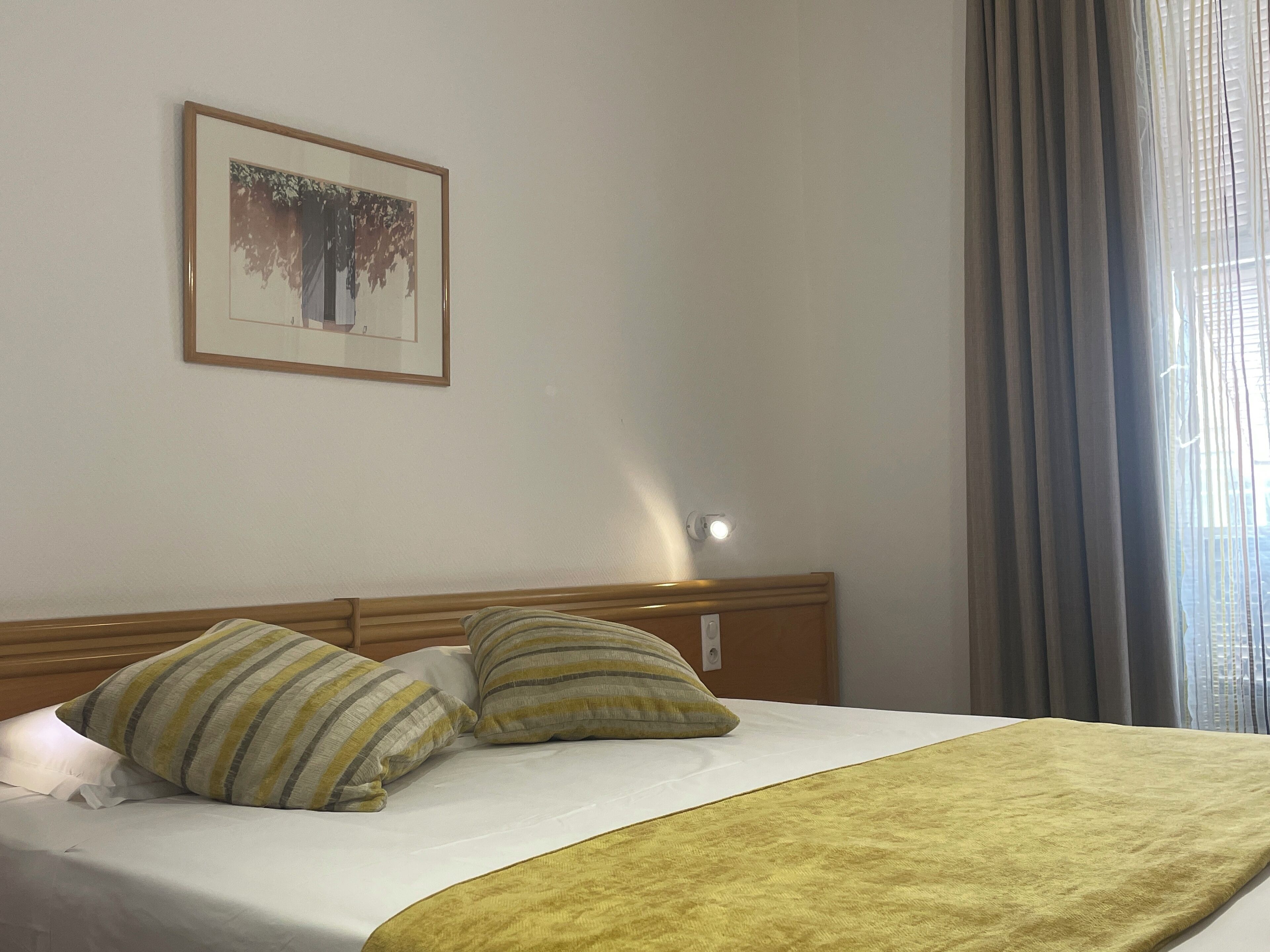 A hotel room with a bed, two pillows, a blanket, a framed picture on the wall, and a window with curtains.