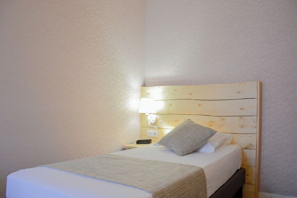A neatly made bed with a wooden headboard, a bedside lamp, and a remote control on a nightstand. - Hotel Busby (Nice)