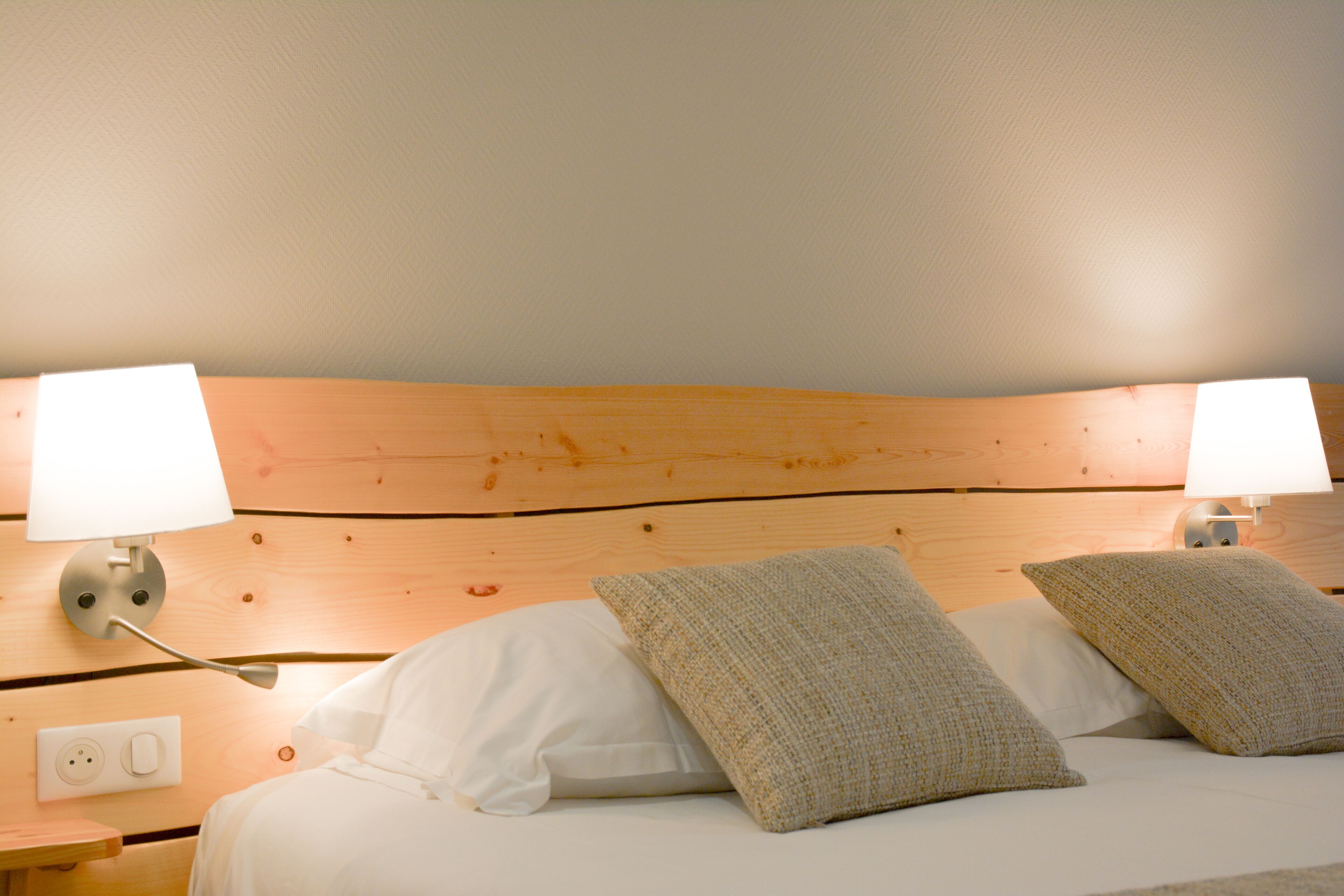Una cama bien hecha con almohadas blancas y con textura, dos lámparas de noche y un cabecero de madera.