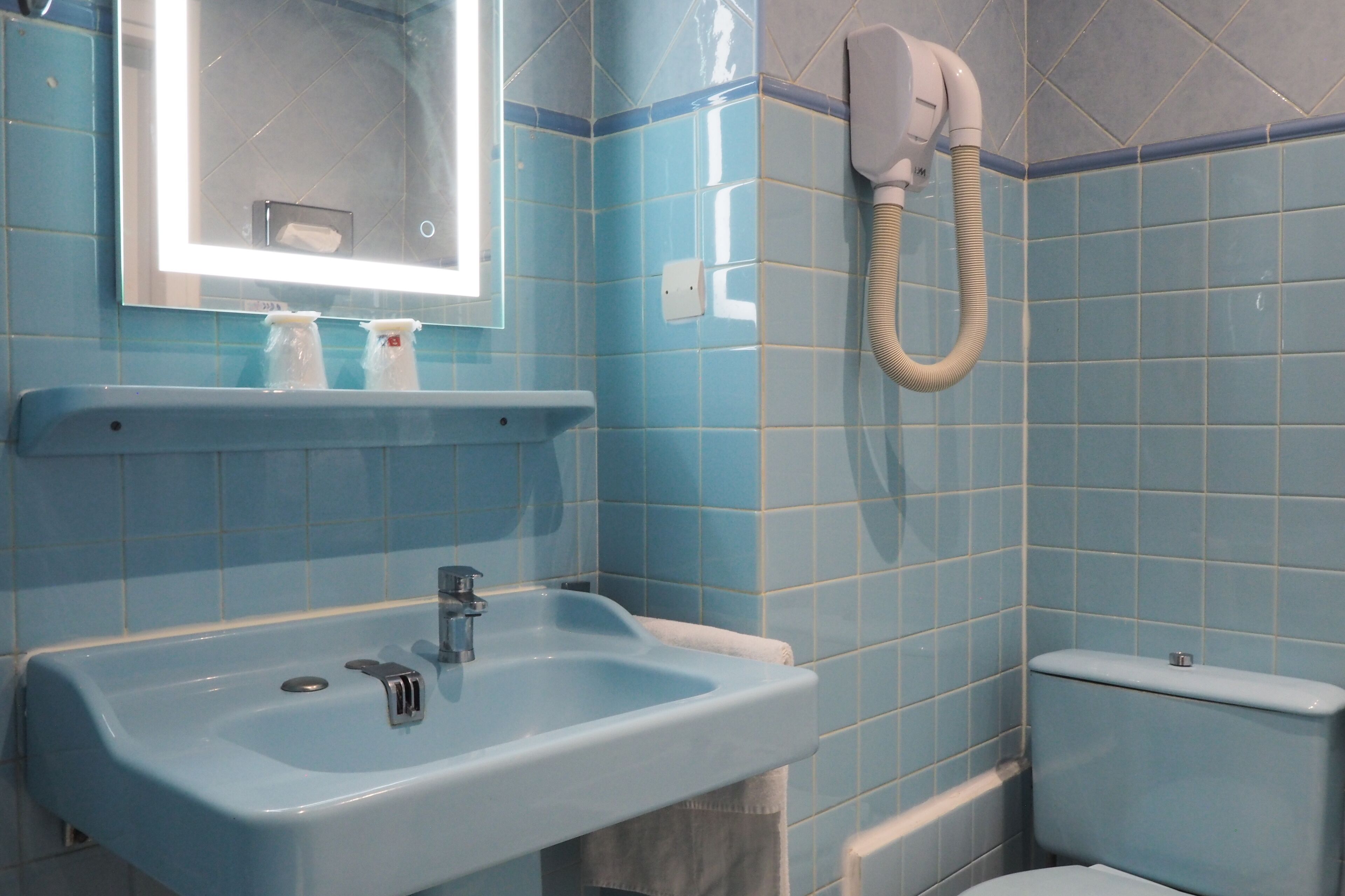 Un baño con pared de azulejos azules, un lavabo con espejo y un inodoro con manguera.