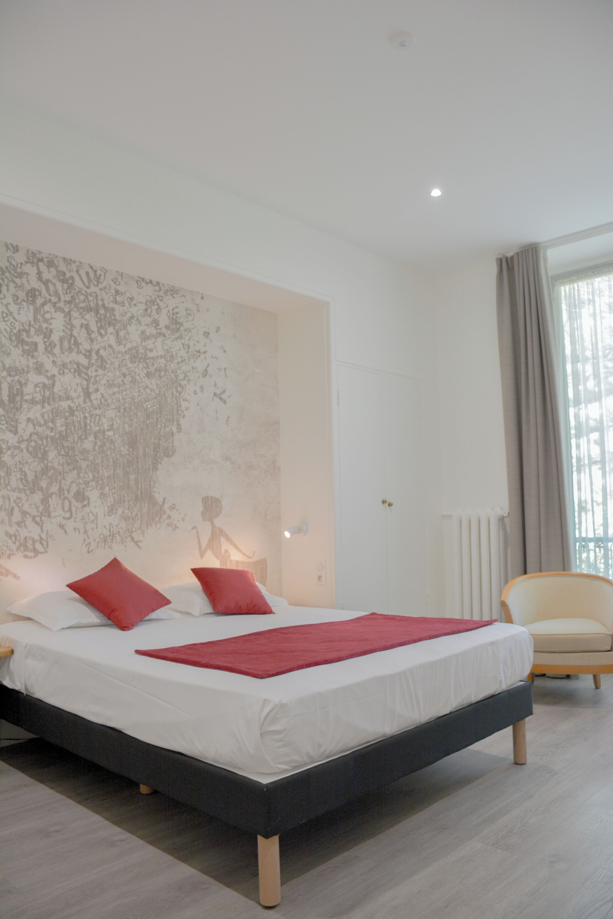 A modern bedroom with a large bed, two red pillows, a bedside table with a lamp, a chair, and a large abstract wall mural.