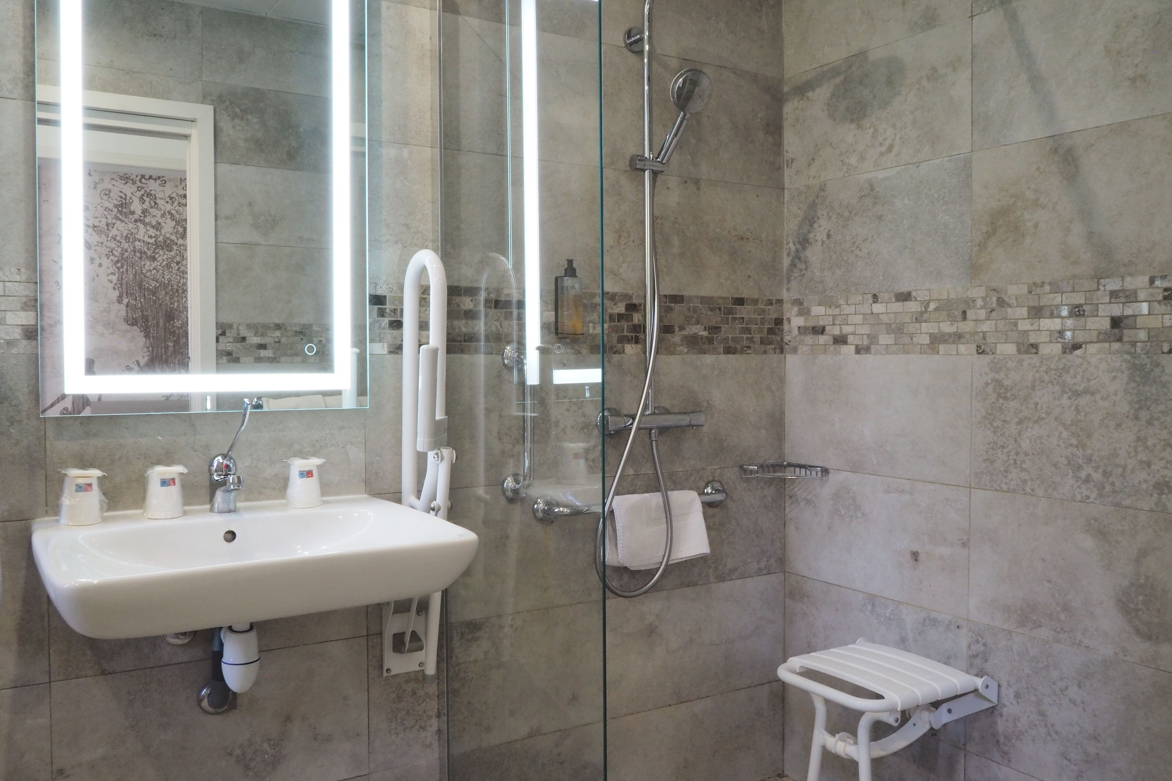 A bathroom with a glass-enclosed shower, a white sink with soap dispensers, and a small white bench.