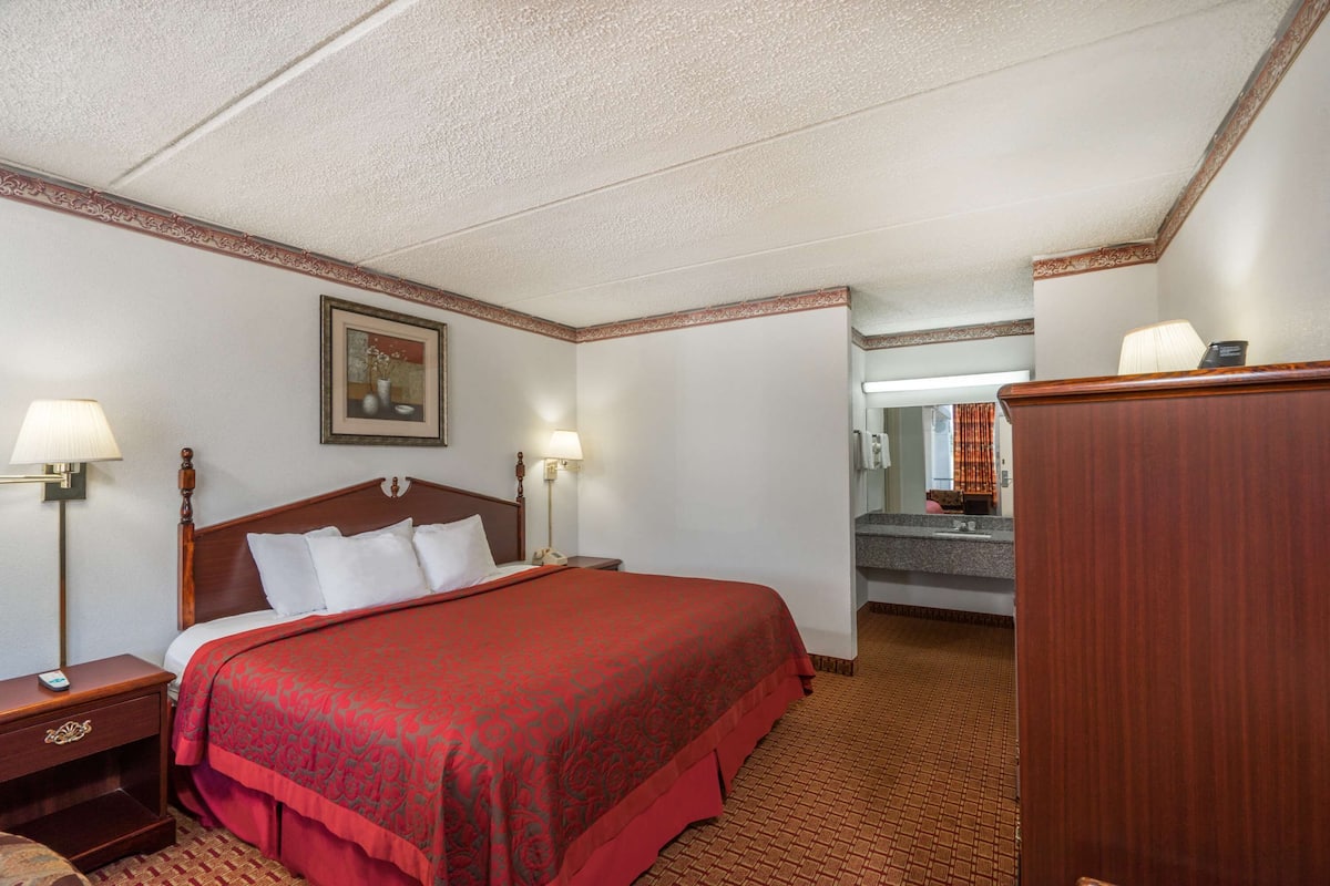A hotel room with a large bed, two bedside lamps, a wooden nightstand, a framed picture on the wall, and a bathroom visible through an open door.